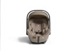 Fotelik samochodowy Joie i-Level Pro w kolorze sandstone (piaskowy beż), widziany od przodu. Fotelik jest w pozycji pionowej, z uniesioną rączką i rozłożonym daszkiem. Zdjęcie przedstawia wkładkę dla niemowlaka oraz pasy bezpieczeństwa.