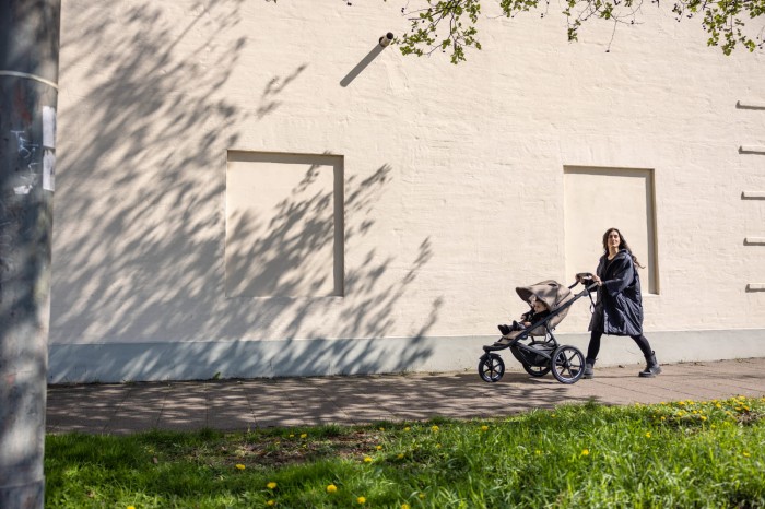 Kobieta prowadzi wózek spacerowy Thule Urban Glide 3 w kolorze Tinted Taupe po miejskim chodniku