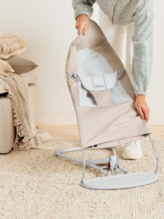 Osoba dorosła składa beżowy leżaczek BabyBjörn Bouncer, opuszczając go na dywan. Prezentuje funkcję łatwego składania i przenoszenia. W tle widać kanapę z poduszkami.