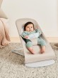 Niemowlę ubrane w miętowy dzianinowy komplet siedzi zabezpieczone pasem w beżowym leżaczku BabyBjörn Bouncer ustawionym na dywanie w pozycji półleżącej.