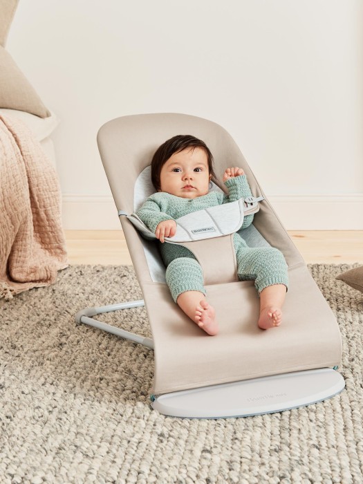 Niemowlę ubrane w miętowy dzianinowy komplet siedzi zabezpieczone pasem w beżowym leżaczku BabyBjörn Bouncer ustawionym na dywanie w pozycji półleżącej.