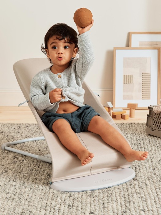 Starsze niemowlę siedzi w beżowym leżaczku BabyBjörn Bouncer (używanym jako fotelik) i podnosi rączkę, trzymając małą piłkę. Leżaczek jest ustawiony w najbardziej pionowej pozycji.