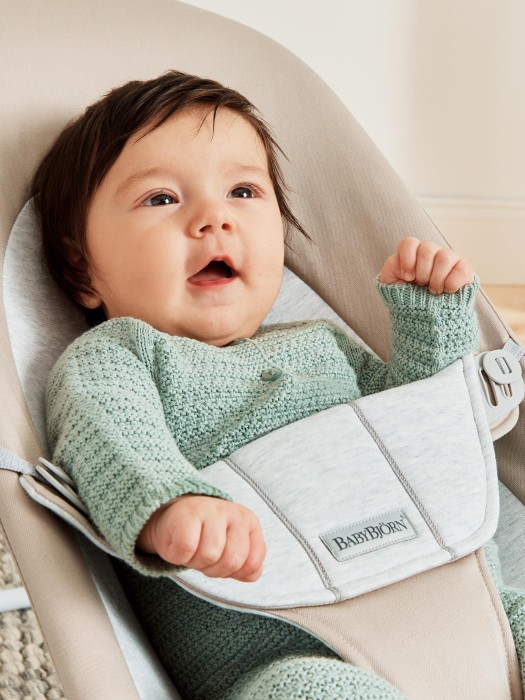 Bardzo bliskie ujęcie uśmiechniętego niemowlęcia w miętowym ubranku, bezpiecznie zapiętego w leżaczku BabyBjörn Bouncer. Eksponuje miękkość siedziska i pas bezpieczeństwa z logo.