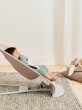 Niemowlę w miętowym ubranku leży w beżowym leżaczku BabyBjörn Bouncer ustawionym w najniższej pozycji. Dziecko obserwuje coś w oddali, obok leżą poduszki i pluszak.