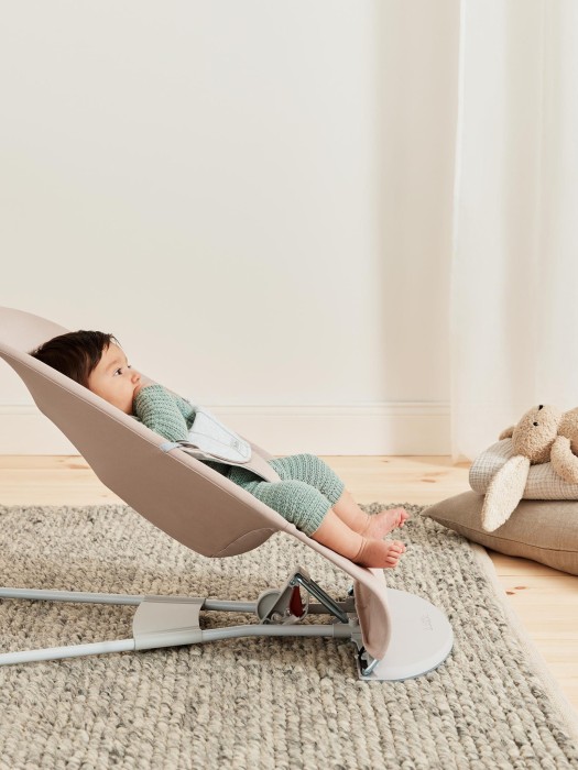 Niemowlę w miętowym ubranku leży w beżowym leżaczku BabyBjörn Bouncer ustawionym w najniższej pozycji. Dziecko obserwuje coś w oddali, obok leżą poduszki i pluszak.