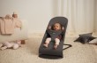 Uśmiechnięte niemowlę leży w szarym leżaczku BabyBjörn na jasnym, futrzanym dywanie w pokoju. Leżaczek jest ustawiony w pozycji półleżącej. W tle widoczne są pluszaki i jasna pufa.
