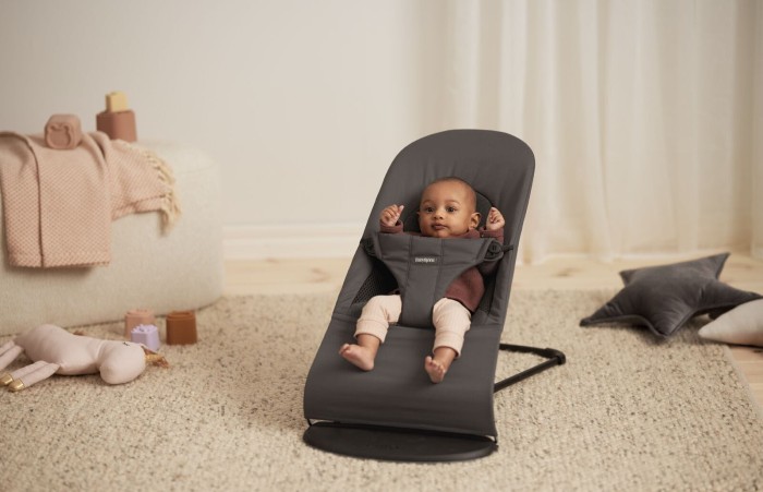 Uśmiechnięte niemowlę leży w szarym leżaczku BabyBjörn na jasnym, futrzanym dywanie w pokoju. Leżaczek jest ustawiony w pozycji półleżącej. W tle widoczne są pluszaki i jasna pufa.