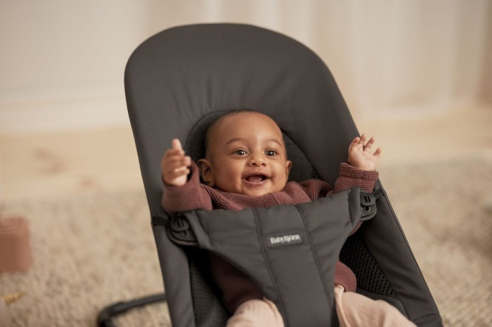 Zbllżenie na radosne, uśmiechnięte niemowlę, które leży w szarym leżaczku BabyBjörn, z uniesionymi rączkami. Widoczne jest logo BabyBjörn na pasach bezpieczeństwa.