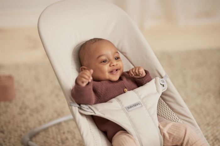 Zbliżenie na radosne, uśmiechnięte niemowlę, które leży w beżowym leżaczku BabyBjörn, z uniesionymi rączkami. Widoczne jest logo BabyBjörn na pasach bezpieczeństwa.