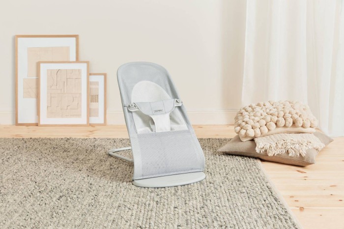 Leżaczek BABYBJÖRN w jasnoszarej siateczce w nowoczesnym, jasnym wnętrzu. Stoi na dywanie, w otoczeniu ramek z grafikami i stosu beżowych poduszek. Leżaczek ustawiony w pozycji pośredniej.