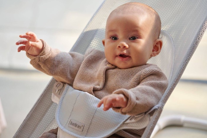 Szczęśliwe niemowlę w beżowym sweterku siedzi w leżaczku BABYBJÖRN MESH, unosząc rączki. Wyraźnie widoczny centralny pas bezpieczeństwa. Ujęcie z bliska, w jasnym świetle, podkreśla radość dziecka.