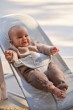 Zbliżenie roześmianego niemowlęcia w dzianinowym ubranku, siedzącego w leżaczku BABYBJÖRN. Widać ergonomiczne oparcie i bezpieczne pasy. Uśmiech dziecka świadczy o zadowoleniu.