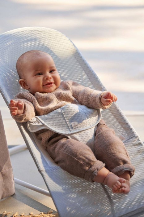 Zbliżenie roześmianego niemowlęcia w dzianinowym ubranku, siedzącego w leżaczku BABYBJÖRN. Widać ergonomiczne oparcie i bezpieczne pasy. Uśmiech dziecka świadczy o zadowoleniu.