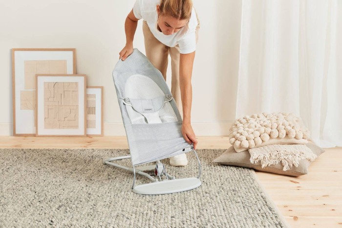 Kobieta w jasnym ubraniu demonstruje składanie leżaczka BABYBJÖRN. Trzyma go pionowo nad dywanem w jasnym wnętrzu, co ilustruje łatwość obsługi i niewielką wagę produktu.
