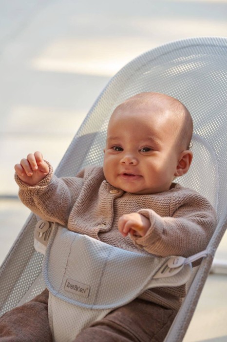 Bliskie ujęcie niemowlęcia w beżowym sweterku, siedzącego w leżaczku BABYBJÖRN. Dziecko patrzy z delikatnym uśmiechem. Kadr skupia się na twarzy i bezpiecznym ułożeniu w leżaczku.