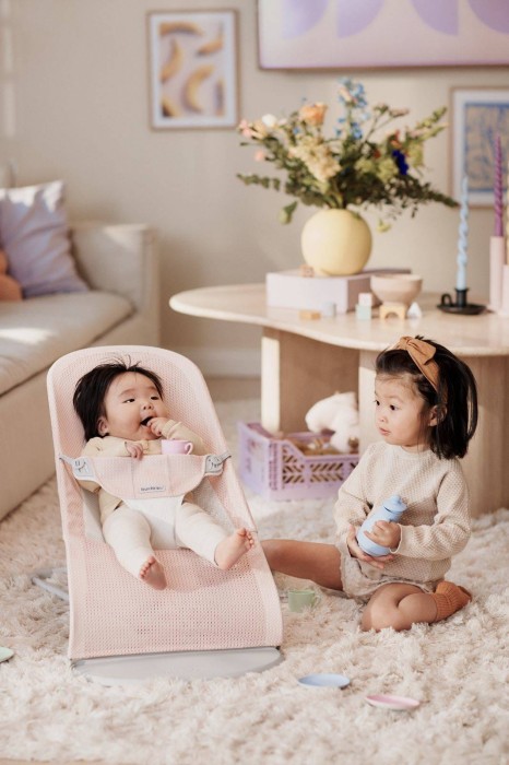 Niemowlę w leżaczku BABYBJÖRN (różowy Mesh) w jasnym salonie. Obok siedzi starsze dziecko, bawiąc się zabawkami. Scena ilustruje codzienne użytkowanie leżaczka w domu.