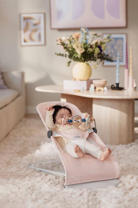 Niemowlę w leżaczku BABYBJÖRN (różowy Mesh) w jasnym salonie, trzymające zabawkę – pałąk z drewnianymi elementami. Dookoła stolik kawowy z wazonem i obrazami w tle.