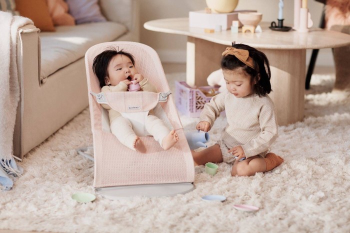 Niemowlę w leżaczku BABYBJÖRN (różowy Mesh) i starsze dziecko bawią się na kudłatym dywanie w salonie. Scena prezentuje leżaczek jako element rodzinnego życia.