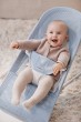 Roześmiane niemowlę w leżaczku BABYBJÖRN (błękitny Mesh), ubrane w beżowy sweterek. Wyraźnie widoczne szerokie rączki i uśmiech dziecka.