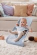Niemowlę w leżaczku BABYBJÖRN (błękitny Mesh) w jasnym salonie, na kudłatym dywanie. Dziecko bawi się, a w tle widać kanapę z kolorowymi poduszkami.