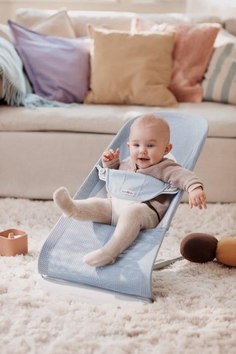 Niemowlę w leżaczku BABYBJÖRN (błękitny Mesh) w jasnym salonie, na kudłatym dywanie. Dziecko bawi się, a w tle widać kanapę z kolorowymi poduszkami.
