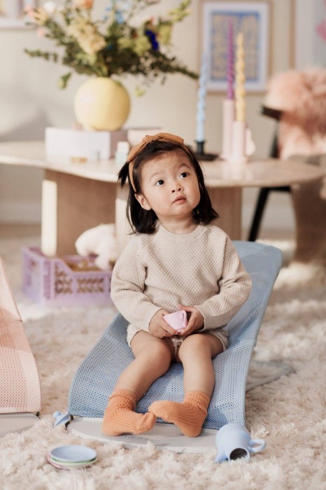 Starsze dziecko siedzi w błękitnym leżaczku BABYBJÖRN (poszycie odwrócone) i patrzy w górę. W tle minimalistyczny salon, co ilustruje funkcję fotelika.