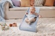 Roześmiane niemowlę siedzi w leżaczku BABYBJÖRN (błękitny Mesh) w jasnym salonie. Wokół zabawki i poduszki. Scena domowa.