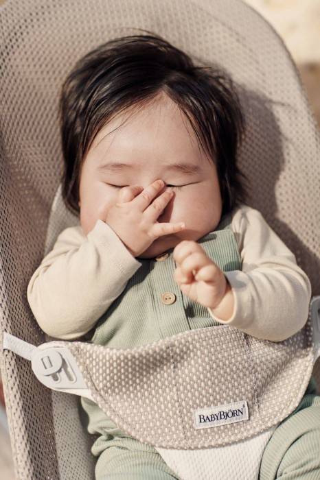 Zbliżenie śpiącego lub drzemiącego niemowlęcia w leżaczku BABYBJÖRN (szarobeżowy Mesh). Rączka dziecka zasłania twarz. Wokół naturalne światło.