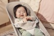 Niemowlę uśmiecha się, leżąc w leżaczku BABYBJÖRN (szarobeżowy Mesh). Kadr z bliska, w jasnym otoczeniu, eksponuje zadowolenie.