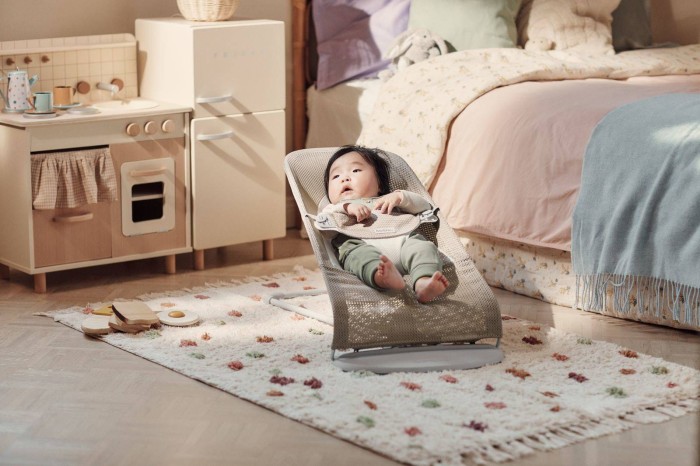 Niemowlę w leżaczku BABYBJÖRN (szarobeżowy Mesh) w jasnym pokoju, obok drewnianej kuchni i łóżka. Dziecko obserwuje otoczenie.