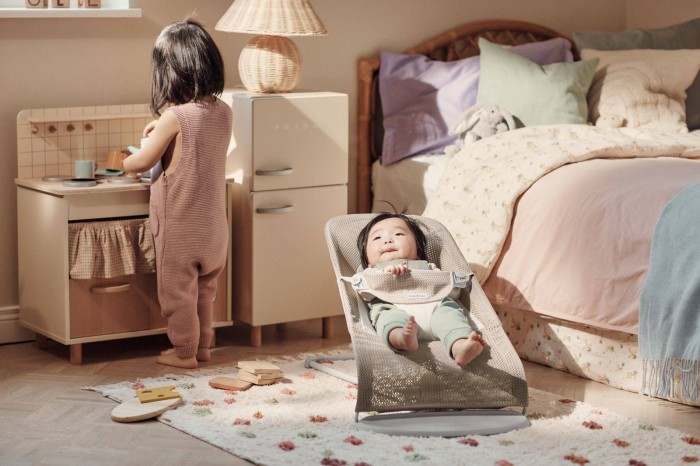 Niemowlę relaksuje się w leżaczku BABYBJÖRN (szarobeżowy Mesh), obserwując starsze dziecko bawiące się przy kuchni. Scena z pokoju dziecięcego.